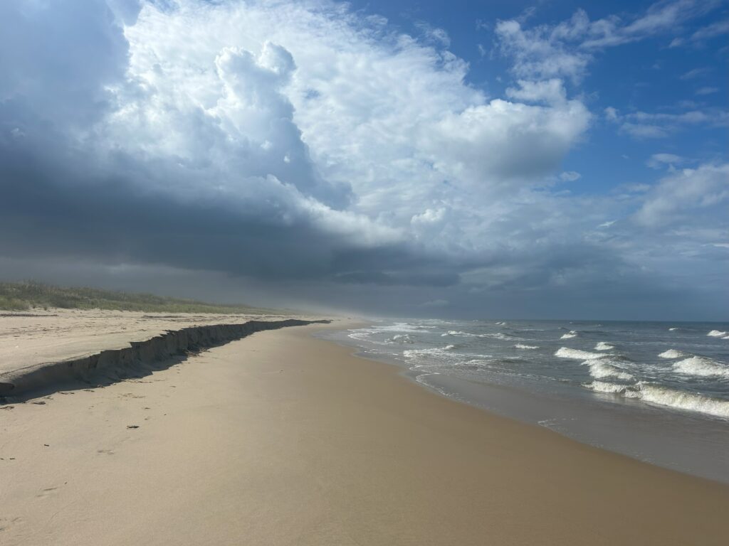 A beach beneath a sky half blue and half gray stormclouds, with a sharp wall of sand that will be eaten by the tide.