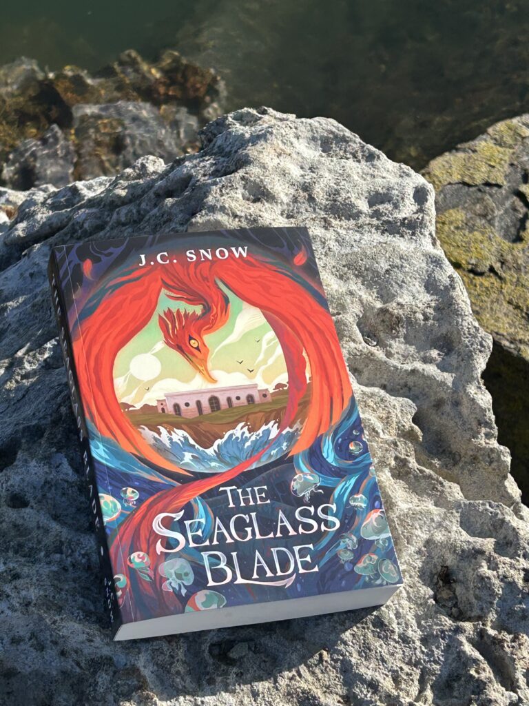 The Seaglass Blade book, lying on a sunny rock overlooking green ocean water.