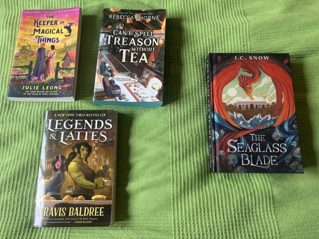 Three books (The Keeper of Magical Things by Julie Leong, Can't Spell Treason without Tea by Rebecca Thorne, and Legends & Lattes by Travis Baldree) are set in a triangle next to a single book, The Seaglass Blade by J.C. Snow.